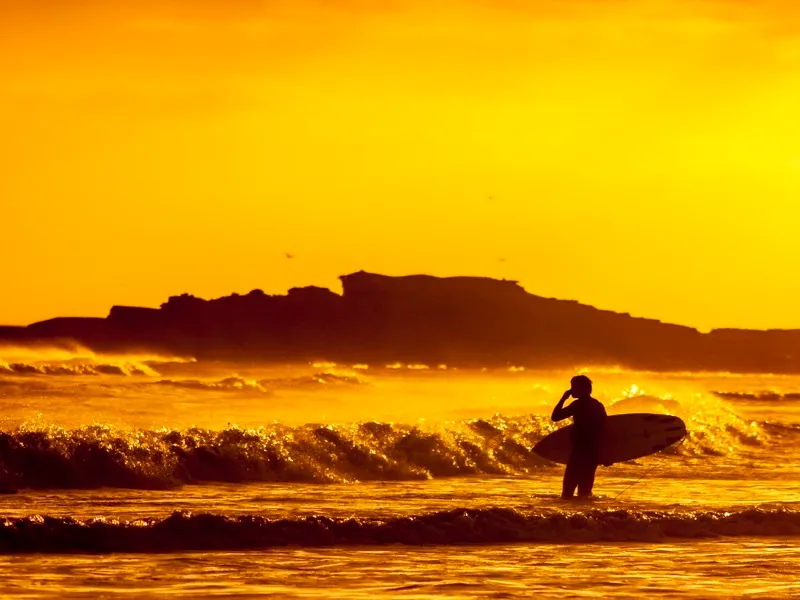 Persona con tabla de surf entrando al mar al atardecer